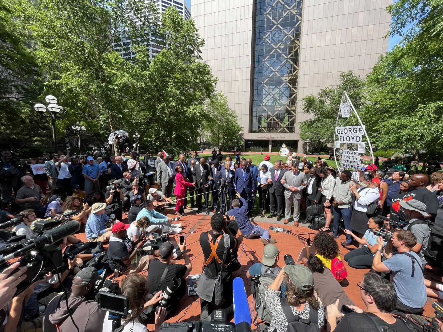 A line of people flank a man speaking into a microphone as reporters with cameras and more microphones look on.