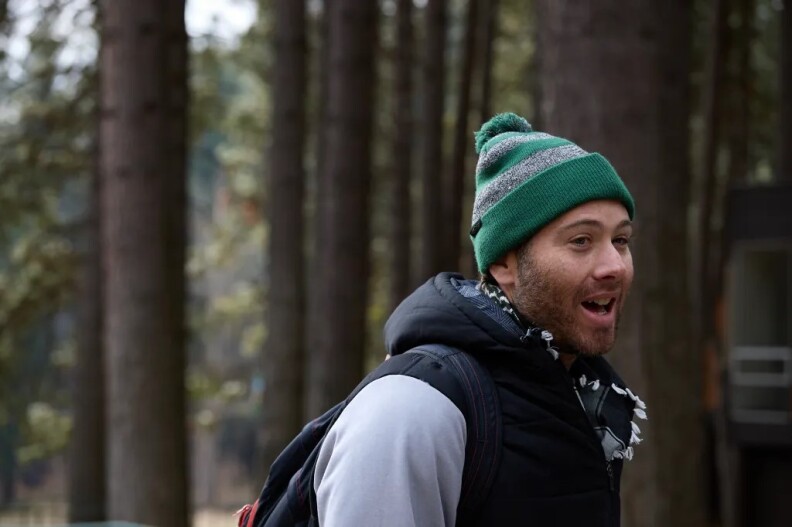 A man wearing a green beanie, dark backpack and black hooded vest