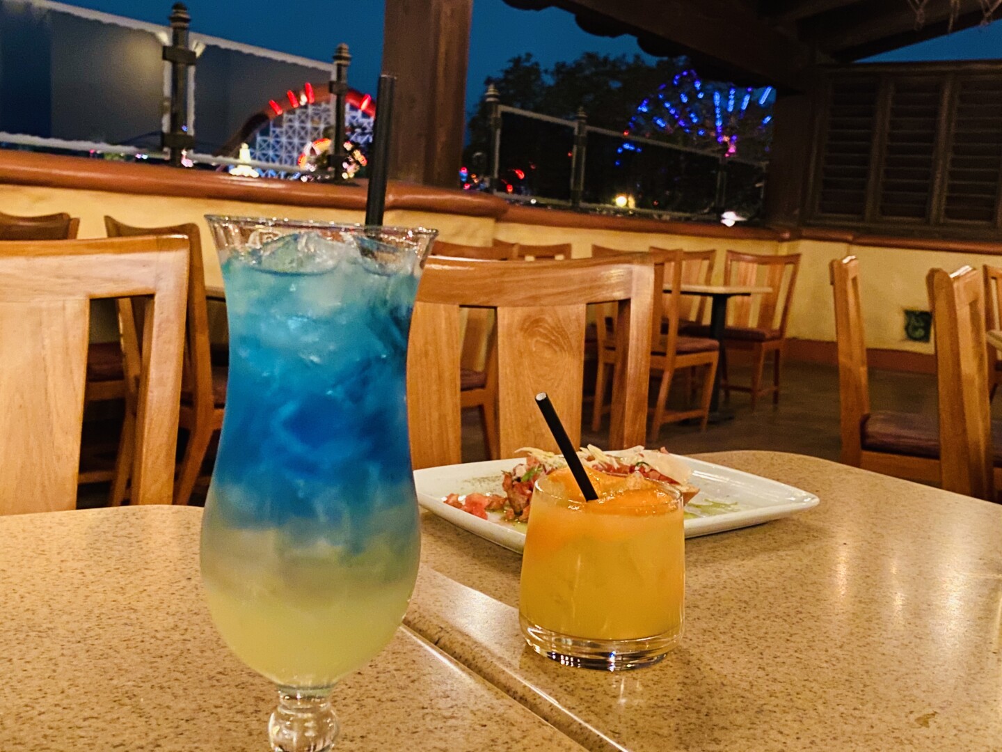 A hurricane glass is filled with blue and yellow liquid. A short, squat glass is filled with pale orange liquid.