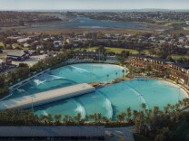 Two pools form a heart shape with a type of pier running down the middle of it: Uniform, generated waves are making their way across the surface. The exterior of the pools are lined with palm trees and building with patios where onlookers can catch the action.  