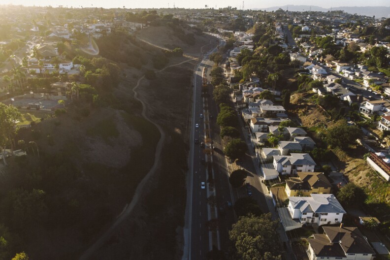 An aerial view shows a long road that splits down the middle of hillsides that have houses on the right and a walking trail surrounded by brush on the left. More houses are seen at the top of the bluff on the left side.