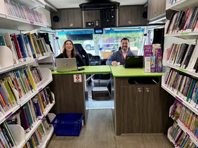 A smiling man and a woman with medium light skin tone sit behind a green counter. There are bookshelves lining either side and a clear windshield behind them. 