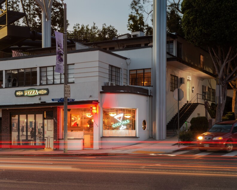 The exterior of a modern Art Deco-style building features a sign on the left that reads 'pizza' and a separate entrance on the right. The right entrance features a small rounded alcove with a neon sign of a martini glass and the words 'cocktails' underneath in its window.