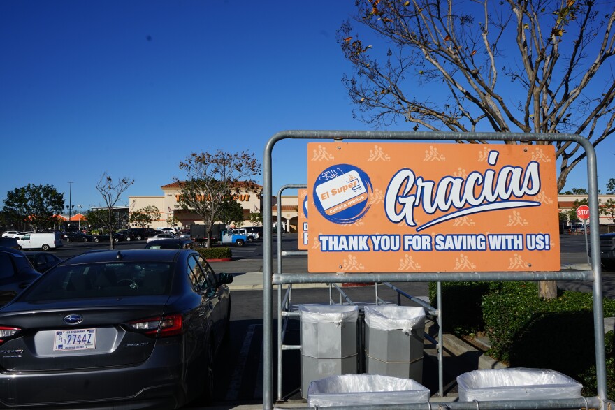An orange sign in a parking lot says "Gracias" in bold white, cursive font. Beneath it, in smaller white font, the sign reads" "Thank you for saving with us!" The sign also has "El Super" branding. Behind the sign is a gray car and a large parking lot in the background.