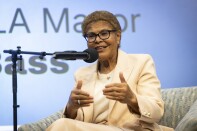 A woman wearing a cream-colored pants suit sits on a chair behind a microphone. 