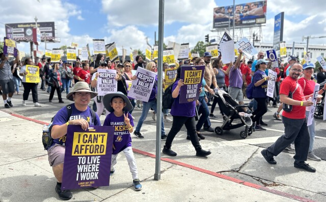 A light-skinned man wearing a tan wide-brimmed hat crouches with on a sidewalk with a sign that reads "I can't afford to live where I work." His young son also wears a wide-brimmed hat and stands to his father's left holding his hands up in a peace sign. They both wear purple shirts. Protestors march in the background.