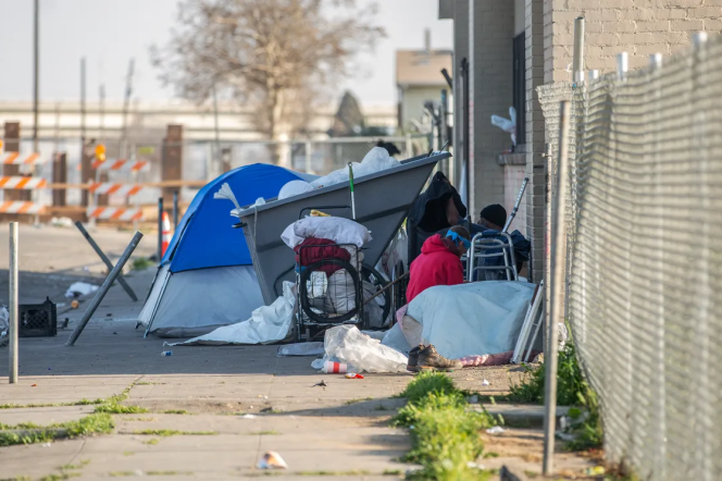 Trash is strewn around an overflowing dumpster on a sidewalk. A blue tent is beside the dumpster.