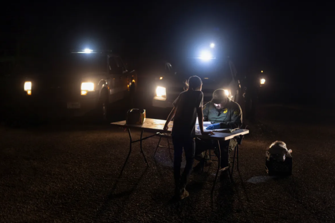 A teenager in a shirt and pnts is leaning over a foldable table and is standing across a pan in a hat and uniform. Two trucks are behind them with their headlights on in the night. 