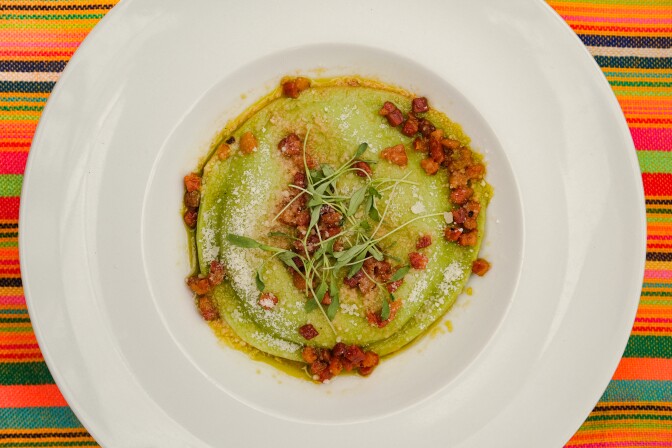 A white plate contains a swirl of green puree, with a red garnish. The plate sits on a multi striped tablecloth with red, green, blue and yellow stripes