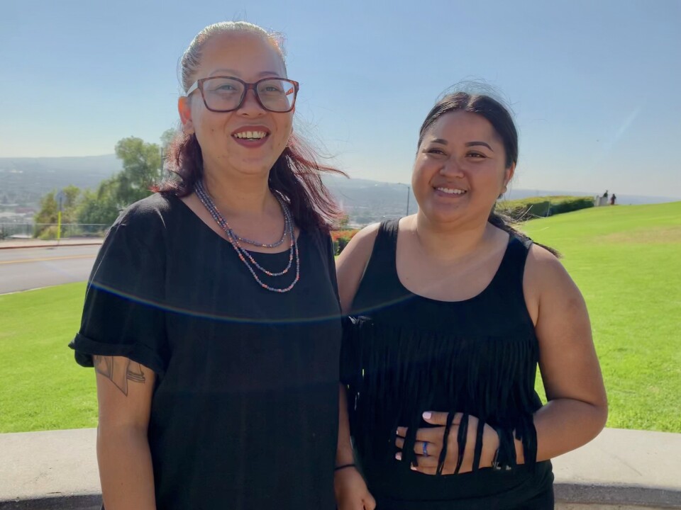 Two women of Asian descent, both wear black, pose together at a park with a harbor as a backdrop.