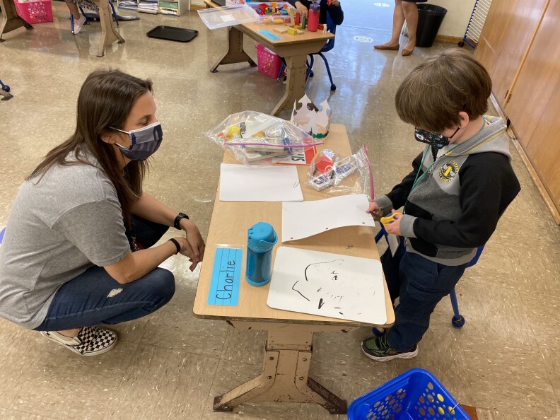 A masked teacher squats down to talk to one of her students, a young boy with brown hair wearing jeans and a sweatshirt and is working on an art project