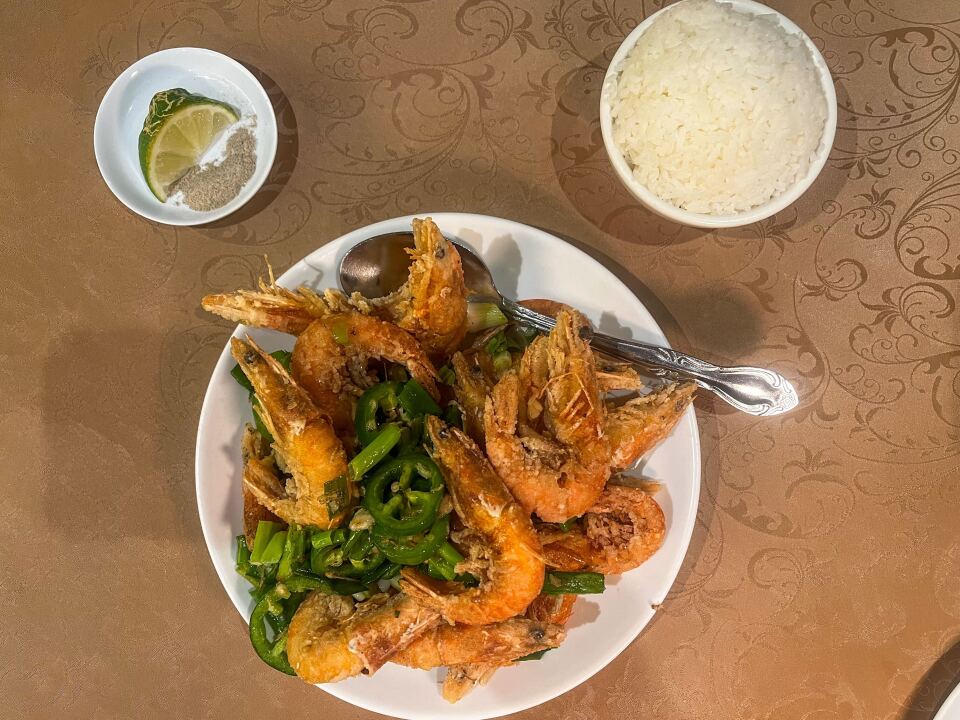 There is a table set with three different dishes of food. On a large white plate at the bottom, there are whole cooked head-on shrimp, a pale orange-pink color. The shrimp are mixed with dark green fresh jalapeño slices and green onion. On the left is a small plate with a slice of lime and a white pepper seasoning. In the right-hand corner is a small bowl of steamed white rice.