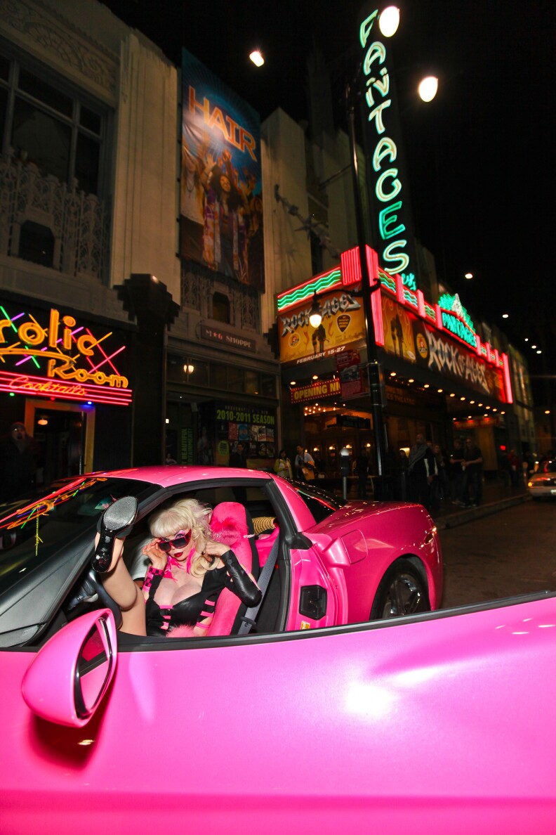 A woman with blond hair and a low-cut top peers over pink sunglasses while sitting in a vibrant pink Corvette.