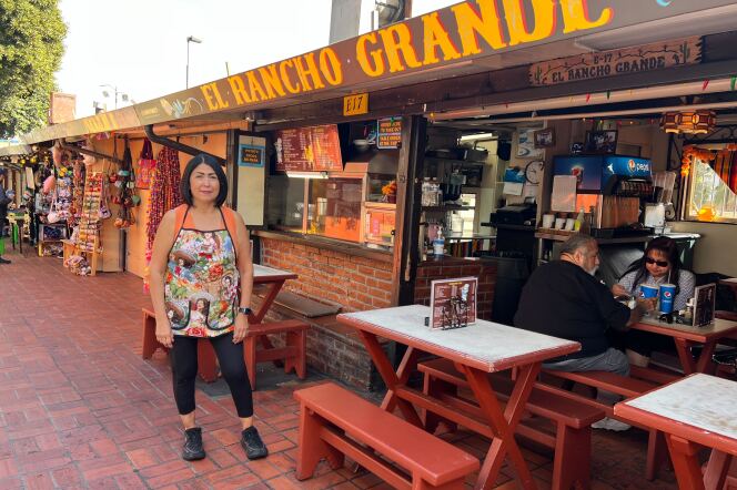 A female presenting person with short black hair wearing a orange sleeveless top and black leggings with a colorful apron stands in front of a restaurant named El Rancho Grande. Picnic style tables are visible. 