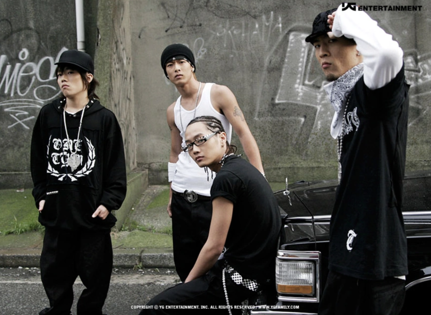 Four Asian men pose for a photo on a street with graffiti behind them. The man on the left wears dark clothes and a black cap. To his right a man wears a white tank top, to his right a man in glasses has his hair in cornrows, and at the far right a man in a cap has his left arm in the air.