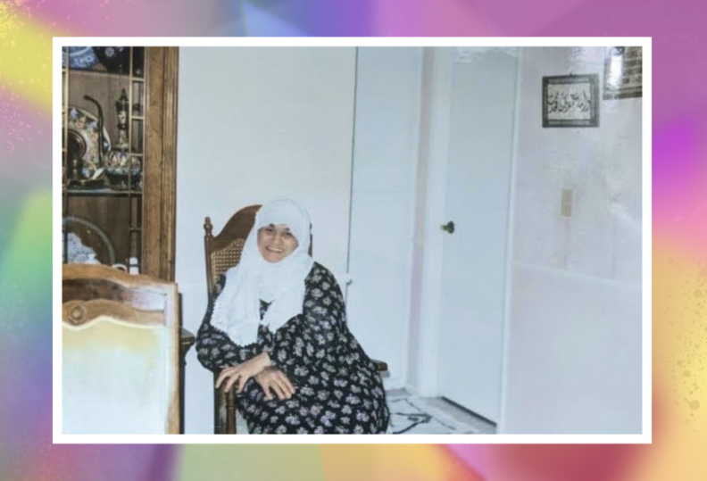 A photo of a smiling woman in a white hijab and black printed dress, seated in a wooden chair.