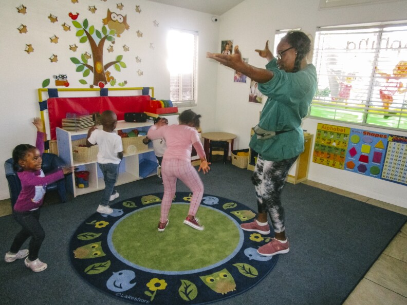 In this image, a teacher and a group of children dance around the classroom.