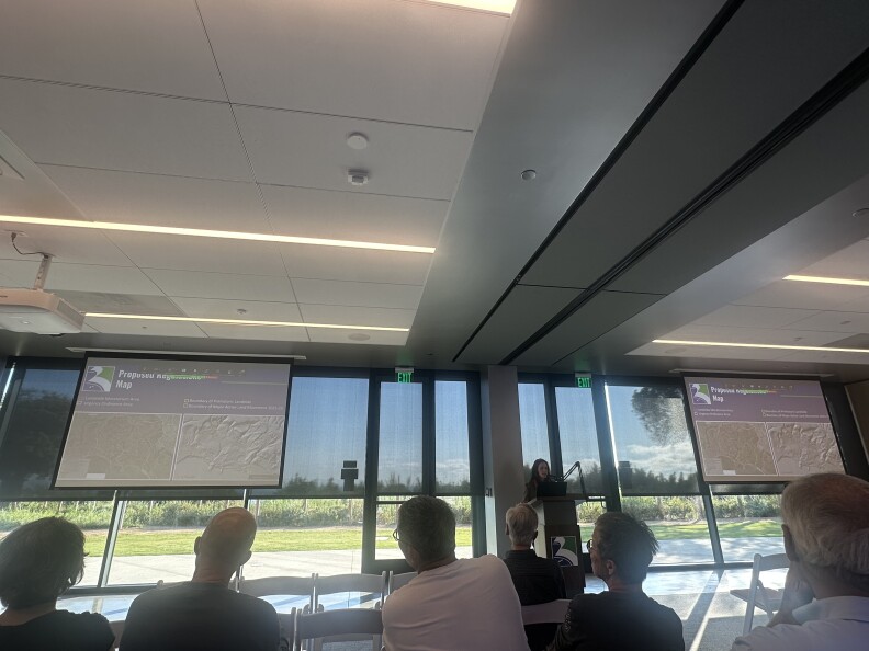 The interior of a community center meeting happening on a sunny day: Many residents are in folding chairs, a person is at a podium and there are two screens at the front of the room.