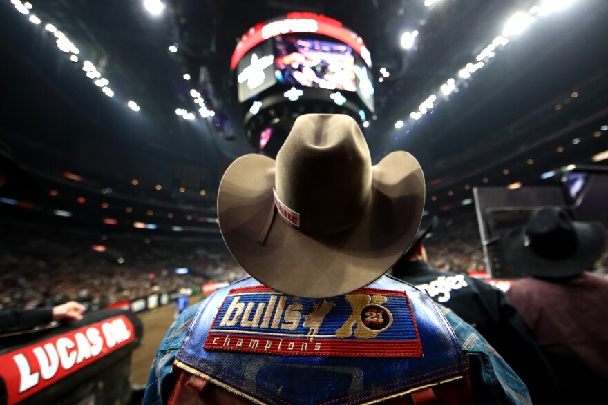 A man wearing a blue jacket with a red and blue patch that reads "bulls 21 champions" wearing a brown curved cowboy hat sits in front of a rodeo arena with dirt on the floor surrounded by blurred dark crowds and an overhead video display.