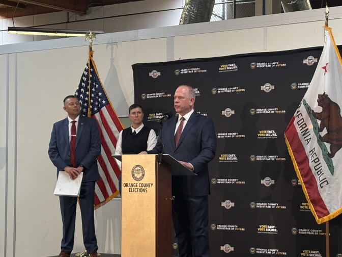 An individual wearing a suit and tie stands at a podium that reads, Orange County elections. There are two other individuals standing nearby on the stage. The stage is flanked by an American flag on the left, and a flag of California on the right. There is a backdrop for the photo that reads "Orange County Registrar of Voters."