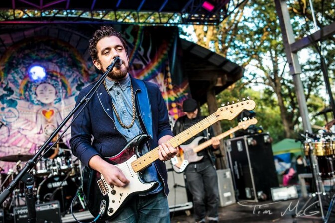A light-skinned man with a beard plays a black and red Stratocaster guitar on a stage. 