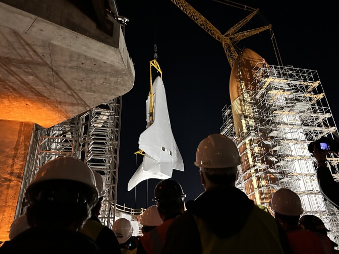 A crowd of people wearing hardhats are looking towards a stark white space shuttle orbiter that's being held vertically in the air by a large yellow crane. The orbiter is near an even-bigger bright orange tank covered in scaffolding. 