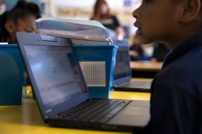 A child seen in profile sits in front of a laptop computer at a communal desk with other students