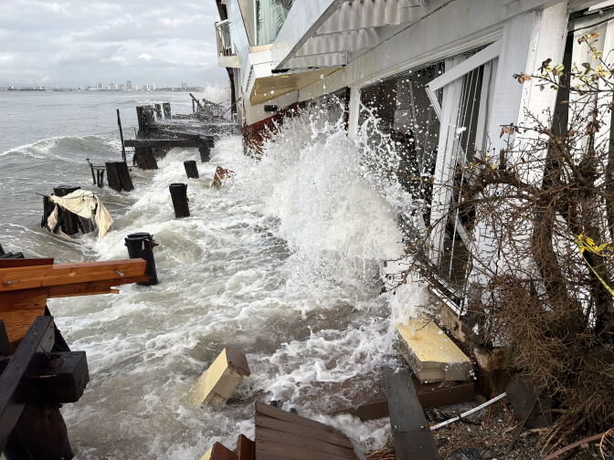 Waves surge against a white waterfront building, splashing up its wall and surrounding broken wooden decking and posts.