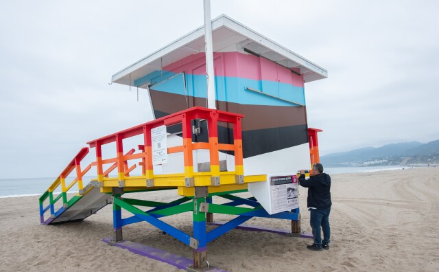 A view of a lifeguard tower on the sand at the beach. It's painted in progress pride flag colors, which are rainbow with the additions of the trans flag colors of white, pink and light blue, and black and brown. A person is near the structure installing part of a sign.
