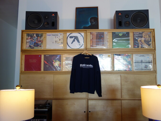 Navy sweatshirt reading “Music works.” hanging on a wooden cabinet that displays two rows of vinyl album covers, with a pair of speakers and a framed painting on top and lamps on either side.