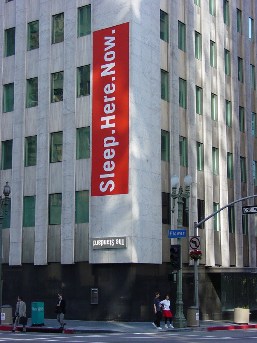 The hotel building with its official upside-down logo "The Standard." A large red sign is on the corner that reads "Sleep. Here. Now."