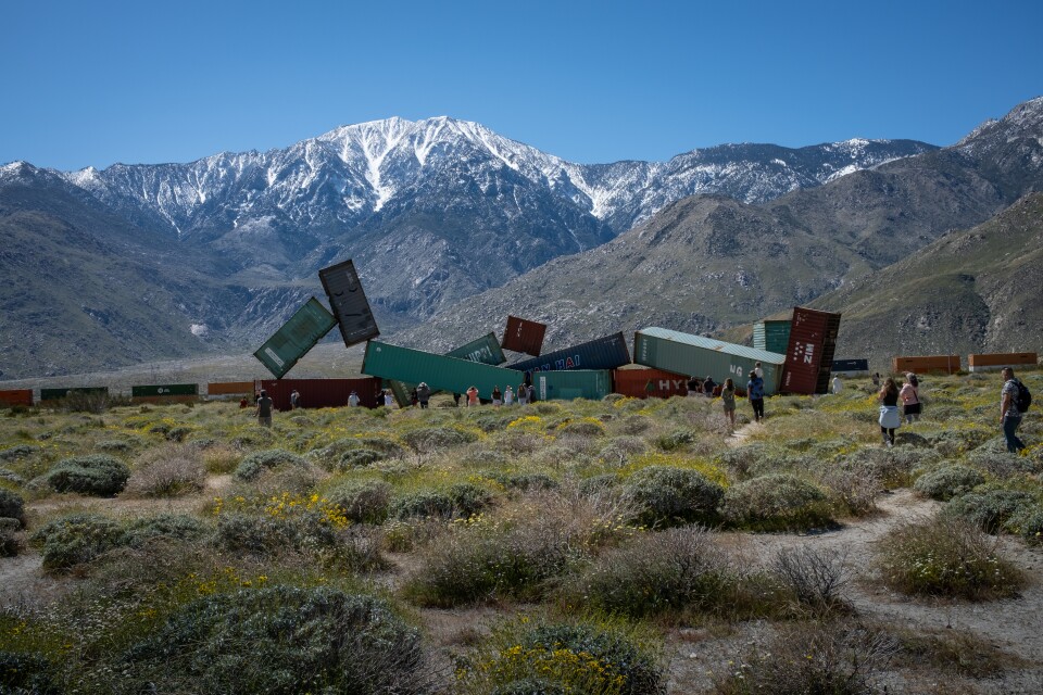 A collection of freight train boxcars assembled to look like a person at rest. There are snow capped mountains behind and wild grasses in the foreground