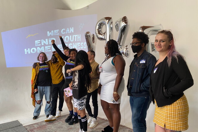 A group of young people stand in front of a crowd, some with the arms pointed up. A screen behind them says we can end youth homelessness.