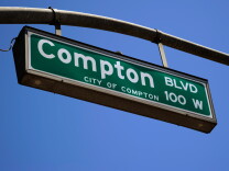 A close-up of a green street sign hanging from a lamp post with a blue sky in the background. The sign reads "Compton Blvd 100 W City of Compton"