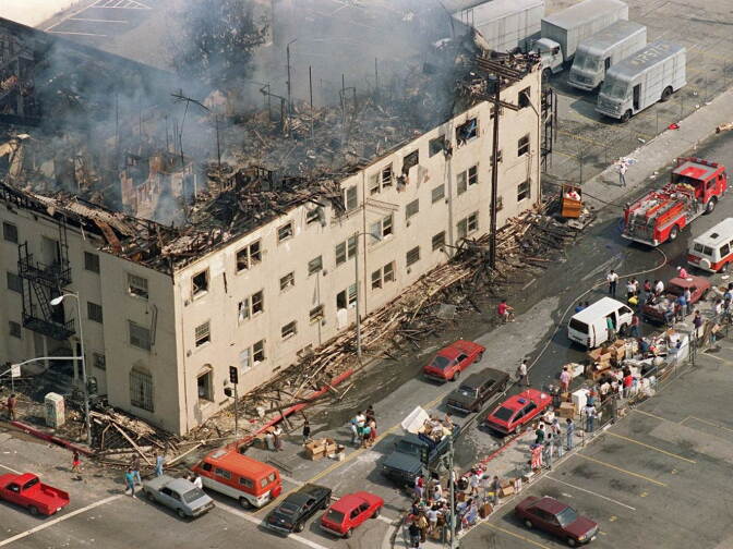 People and their belongings line a sidewalk across from a burned out apartment in Los Angeles. The apartment was attached to a row of stores that were burned and looted, 30 April 1992. 

