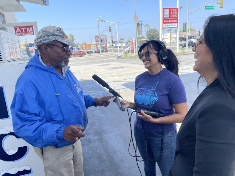 A Black man in a blue jacket with a camouflage cap is interviewed with a mic by an Indian woman wearing headphones, next to an Asian with a black blazer and sunglasses.