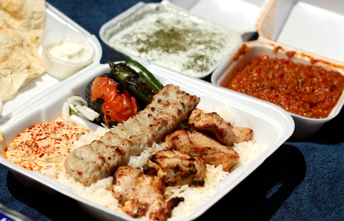 a styrofoam container holds ground meat, a kabob, rice and hummus. other containers hold a red paste and a white yogurt sauce.