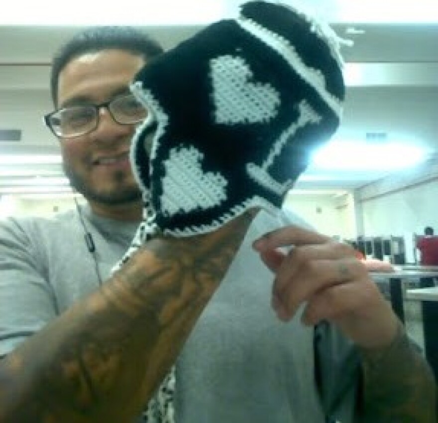 A bearded man in a t-shirt and glasses smiles as he holds up a crocheted black-and-white cap with two hearts on the side.