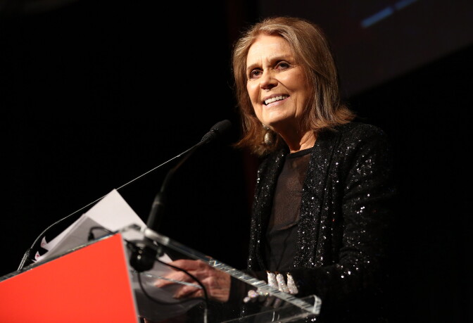 A ssmiling woman wearing a sequined jacket stands at a podium indoor. Behind her is a pitch black background.