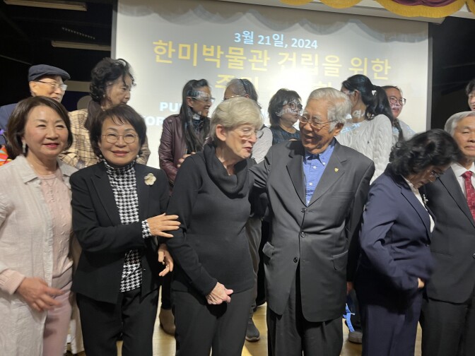 Fourteen people of varying ages, most of them of Korean descent, pose for photos on a stage.