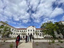 A restored historical building with two visible floors is framed by clouds above and people walking in the foreground.