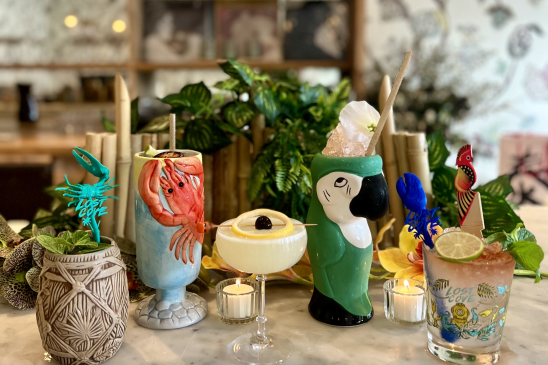 Five tiki-themed cocktails are displayed on a table in Le Fe bar. One of the glasses features a red lobster. Another is made in the shape of a green parrot.