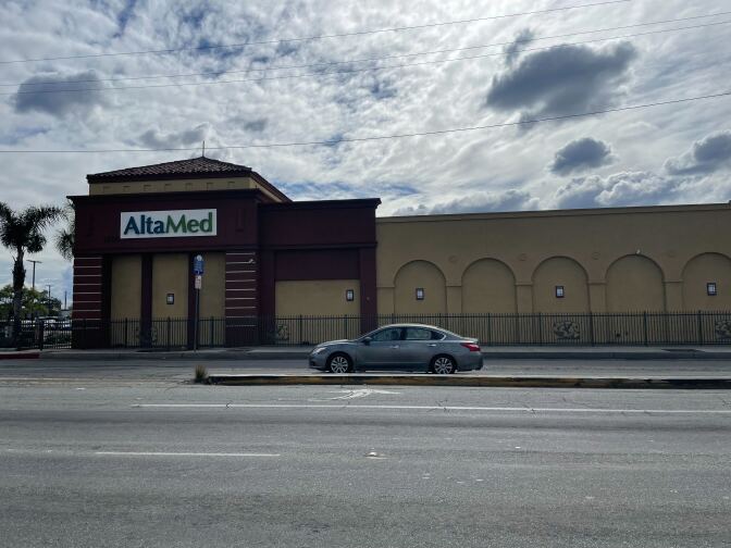 A wide street, empty except for one gray car, in front of a tan building with brick portions on its corner. The building has a sign reading "AltaMed." There is a sunny sky above, but largely covered by fluffy white clouds.