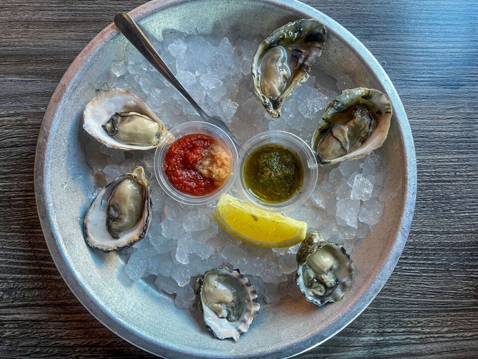 A round metal bowl contains six shucked oysters on crushed ice, arranged in a circle. In the center are two plastic containers with a red sauce on the left and a green one on the right. Next to them is a wedge of lemon.