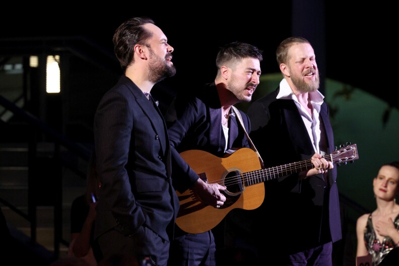 Ben Lovett, Marcus Mumford, and Ted Dwane of the band Mumford & Sons sing together on stage. Marcus, in the middle, is playing acoustic guitar. In the bottom right of the photo, a woman in the audience can be seen watching them, holding a hand to her chest.