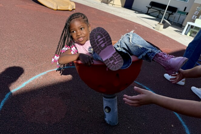  A student with dark skin tone and brown hair in braids with pink beads spins in a chair-like toy on the playground.