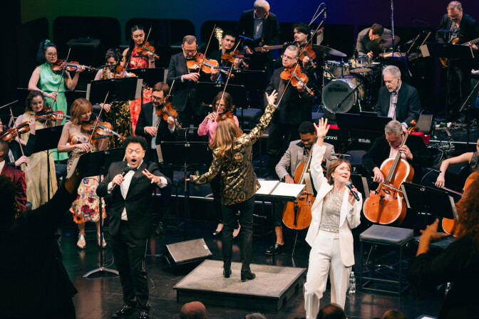 A man in a black tuxedo and a woman in a white suit sing while an orchestra plays behind them. 