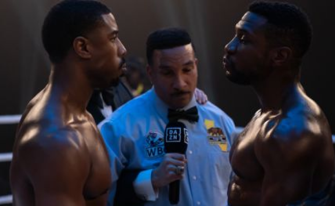 Michael B. Jordan and Johnathan Majors stand in a boxing ring. The room is dark around them except for a bright spotlight that illuminates the center of the ring. Jordan is on the left facing Majors who is on the right. Between them is a referee in a light blue button up with a black bow-tie. He is holding a microphone and looking down at the ground as the other two men stare each other down. Both Majors and Jordan are shirtless, their skin glistening with sweat. 