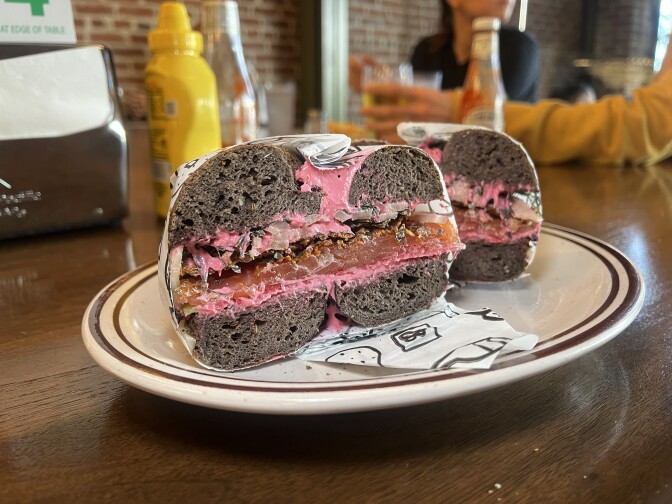 A brown bagel sandwich cut down the middle reveals its contents. It is made of pink cream cheese, chopped white onion, and a thick dark red piece of smoked salmon in the center. The sandwich, wrapped in white to-go paper, sits on a white plate with a brown rim around the edges on a dark brown wood countertop. 