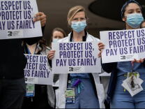 Resident Assistants in blue scrubs and light blue masks hold up white signs that say in purple letters, "Respect us, Protect us, Pay us fairly." the resident in the middle of the photo has blonde hair and looks directly into camera behind wire-framed glasses. There a few other physicians behind them.